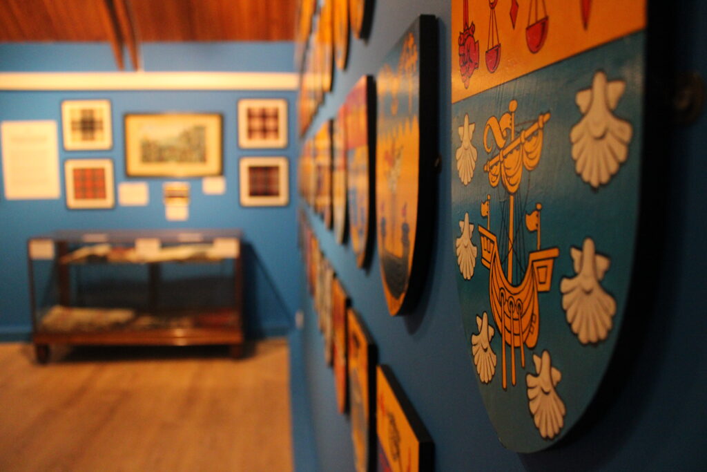 Heraldic shields display inside the Clan Macpherson Museum, with one shield in focus against blue walls and framed tartans.