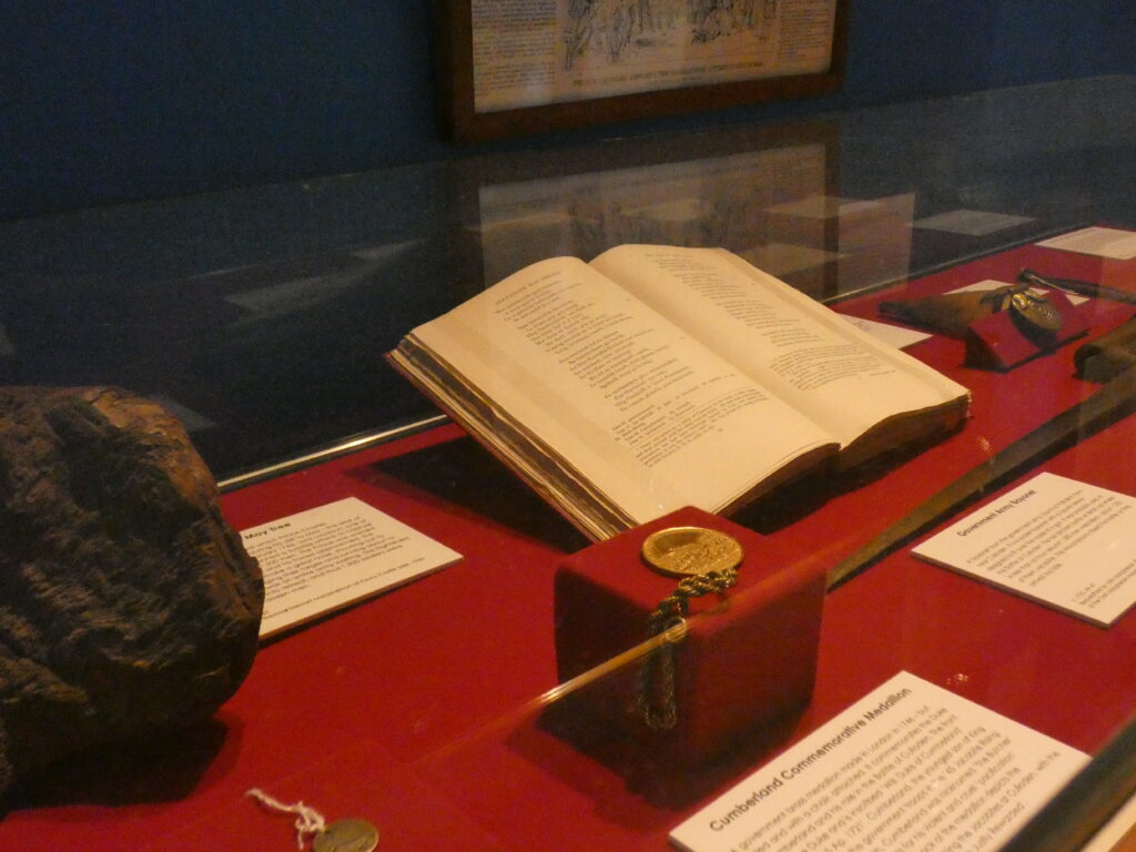 Interior: Exhibition Case Featuring Book and Medallion Museum display case with book, medallion, and Jacobite artefacts on red fabric.