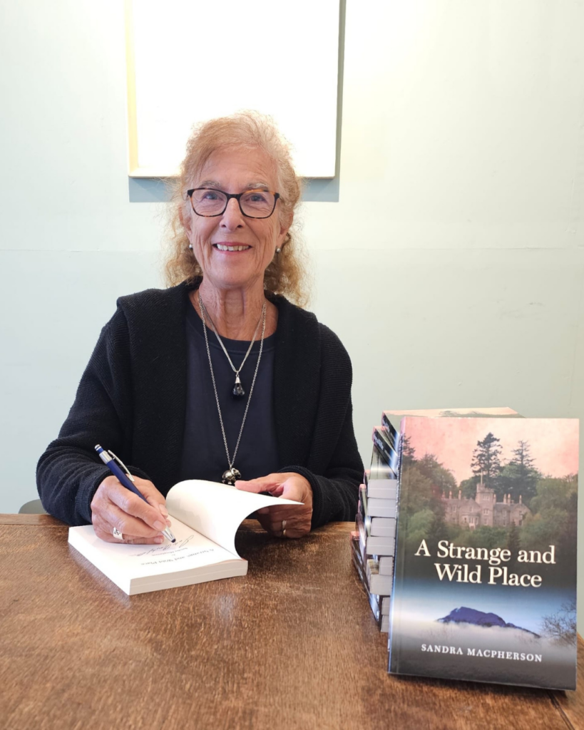 Sandra Macpherson signing a copy of her book, A Strange and Wild Place, with a stack of books beside her.